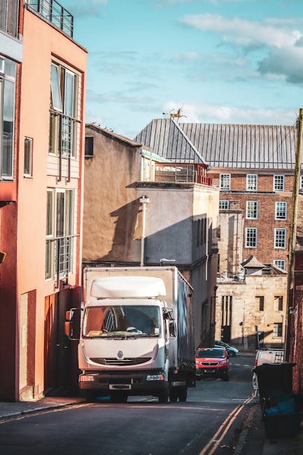 A white box truck parked on a narrow, inclined street in Ickenham, with part of the van close to a multi-story residential building with peach-colored walls and large windows. The area is shaded by surrounding older brick and concrete buildings with flat and slightly sloped roofs. The street features double yellow lines and is lined with various vehicles, including a small red car and a black trash bin. In the background, the sky is partly cloudy with patches of blue. This scene illustrates a typical home relocation or furniture transport scenario within a confined urban street, highlighting access restrictions that specialists like Man and Van Ickenham address during removals and packing and moving operations.