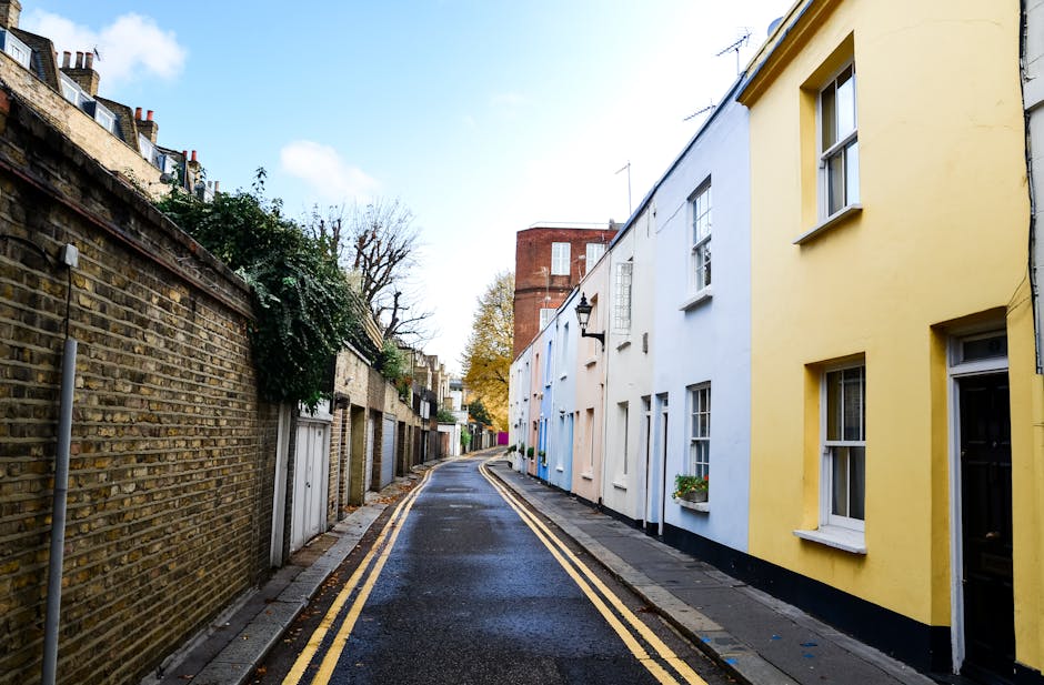 A narrow residential street in Ickenham featuring a pavement on the right side and a brick wall on the left, with the street surface appearing wet and marked with double yellow lines. On the right, there are two-story terraced houses painted in pastel shades of yellow, blue, and white, each with white-framed windows and small flower boxes beneath some of them. A traditional black wall-mounted lantern is attached to one house. In the background, a red brick building is visible along with leafless trees, suggesting late autumn or early winter. The scene is illuminated by natural daylight under a partly cloudy sky. This setting illustrates urban house removals or furniture transport in tight street environments, highlighting the importance of careful planning during home relocation processes. Occasionally, Man and Van Ickenham’s services may be referenced for managing such logistics in challenging street layouts.
