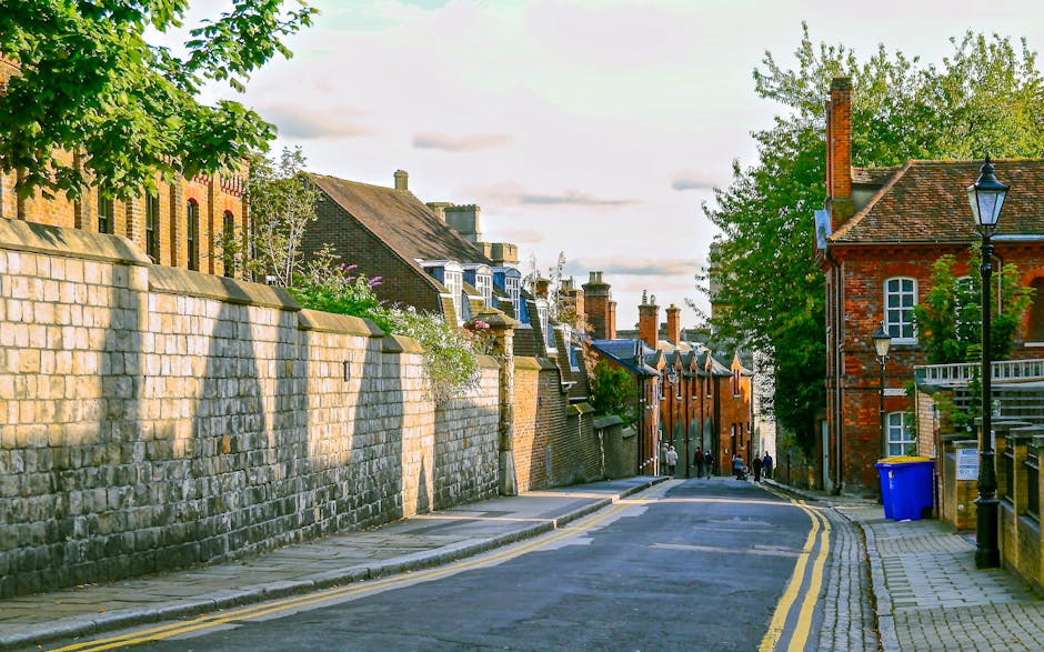A white box truck parked on a narrow, inclined street in Ickenham, with part of the van close to a multi-story residential building with peach-colored walls and large windows. The area is shaded by surrounding older brick and concrete buildings with flat and slightly sloped roofs. The street features double yellow lines and is lined with various vehicles, including a small red car and a black trash bin. In the background, the sky is partly cloudy with patches of blue. This scene illustrates a typical home relocation or furniture transport scenario within a confined urban street, highlighting access restrictions that specialists like Man and Van Ickenham address during removals and packing and moving operations.