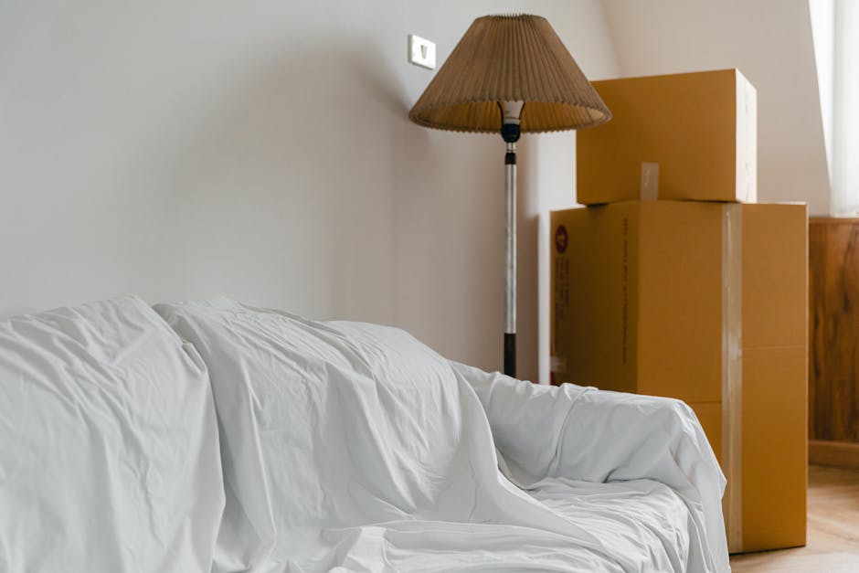 A living room interior showing a white fabric-covered sofa on the left side, partially draped over with crumpled cloth, indicating packing or moving preparation. Behind the sofa, two cardboard moving boxes of different sizes are stacked vertically near a wooden piece of furniture, likely a cabinet or sideboard. A tall floor lamp with a beige fabric lampshade and a metallic stand stands adjacent to the boxes, providing ambient lighting. The boxes are sealed with clear packing tape and are positioned near the corner of the room, close to a window allowing natural light to illuminate the scene. The setting suggests a home in the process of packing or unpacking during a house relocation or move, with visible furniture and packing materials arranged in a typical moving environment. Man and Van Ickenham provides professional removals and furniture transport services, supporting smooth home relocation logistics.