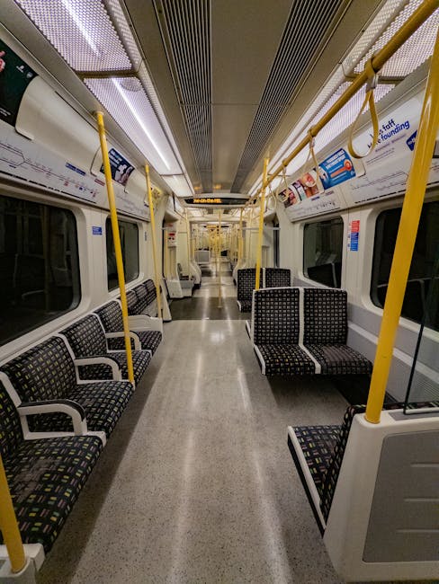 Interior view of an empty city train carriage with rows of black and multicoloured patterned fabric seats along both sides. The carriage has a grey, speckled floor and yellow vertical handrails, with yellow overhead poles and hand straps for standing passengers. The ceiling features a metallic, ribbed design with integrated lighting panels. Large, rectangular windows with black frames are visible on each side, some with advertisements and safety notices. The space appears clean and well-maintained, with no passengers present, capturing a calm scene of urban public transportation, which could be relevant to home relocation or moving logistics when considering transport options for furniture and packing materials. Man and Van Ickenham occasionally operates in local areas, providing removals and furniture transport services.
