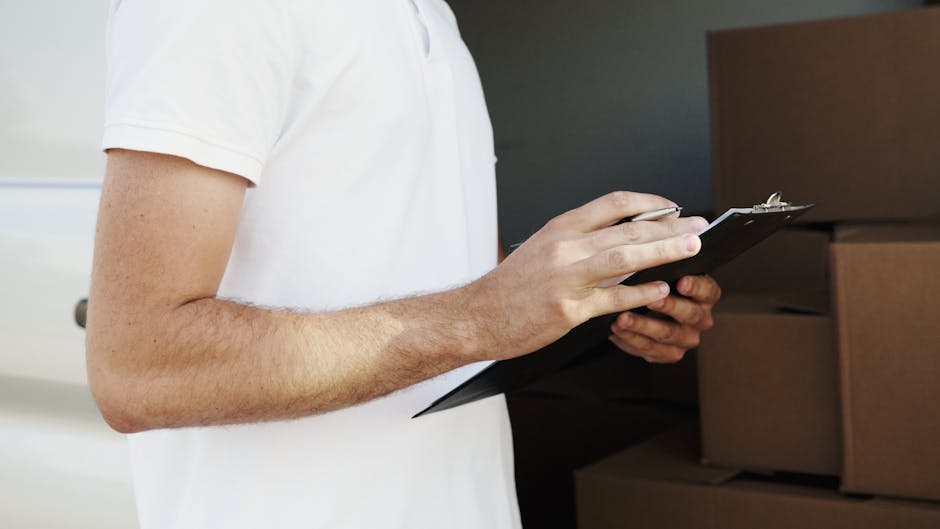 Close-up of a person wearing a white t-shirt holding a black clipboard with a pen, standing beside stacked cardboard moving boxes inside a property. The boxes are sealed with packing tape and vary in size, with some stacked on top of each other near the doorway or room entrance. The individual's arm is visible, showing light skin and some arm hair, while the background includes dark patterned walls and a white door frame. This scene depicts the planning and organisation stage of home relocation or furniture transport, with the person possibly checking inventory or confirming moving details during the packing and loading process. The environment is well-lit, emphasizing the actual objects and their positions, which aligns with professional removals and house moving services offered by Man and Van Ickenham, supporting effective moving logistics preparation.