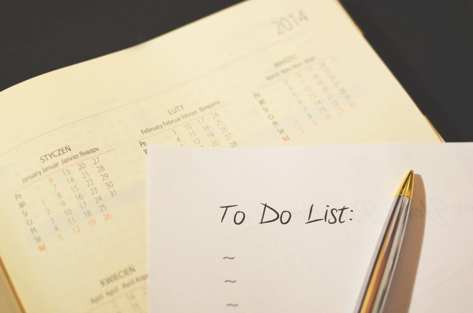 A close-up view of a 2014 calendar page showing months of January through July, with dates marked in black, red, and orange ink. The calendar is laid flat on a dark surface, with the months printed in multiple languages. In front of the calendar, there is a white piece of paper titled 'To Do List:' handwritten in black ink, with several blank lines beneath it for tasks. A gold and silver ballpoint pen rests partially on the paper, angled toward the right. The scene is well-lit, highlighting the details of the calendar, the paper, and the pen, illustrating a workspace setup for planning and organizing house removals or moving logistics, emphasizing the importance of scheduling and task management in home relocation processes, as handled by companies like Man and Van Ickenham.