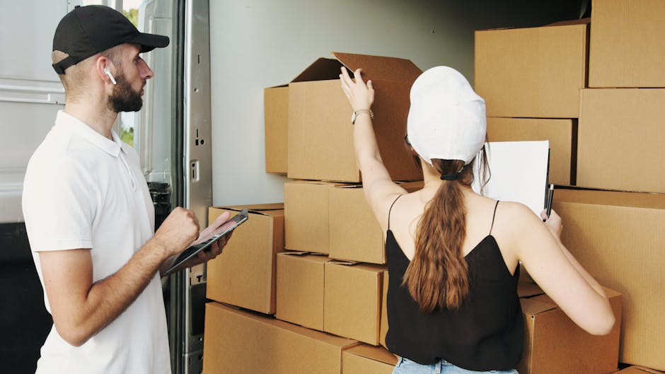 Close-up of a person wearing a white t-shirt holding a black clipboard with a pen, standing beside stacked cardboard moving boxes inside a property. The boxes are sealed with packing tape and vary in size, with some stacked on top of each other near the doorway or room entrance. The individual's arm is visible, showing light skin and some arm hair, while the background includes dark patterned walls and a white door frame. This scene depicts the planning and organisation stage of home relocation or furniture transport, with the person possibly checking inventory or confirming moving details during the packing and loading process. The environment is well-lit, emphasizing the actual objects and their positions, which aligns with professional removals and house moving services offered by Man and Van Ickenham, supporting effective moving logistics preparation.