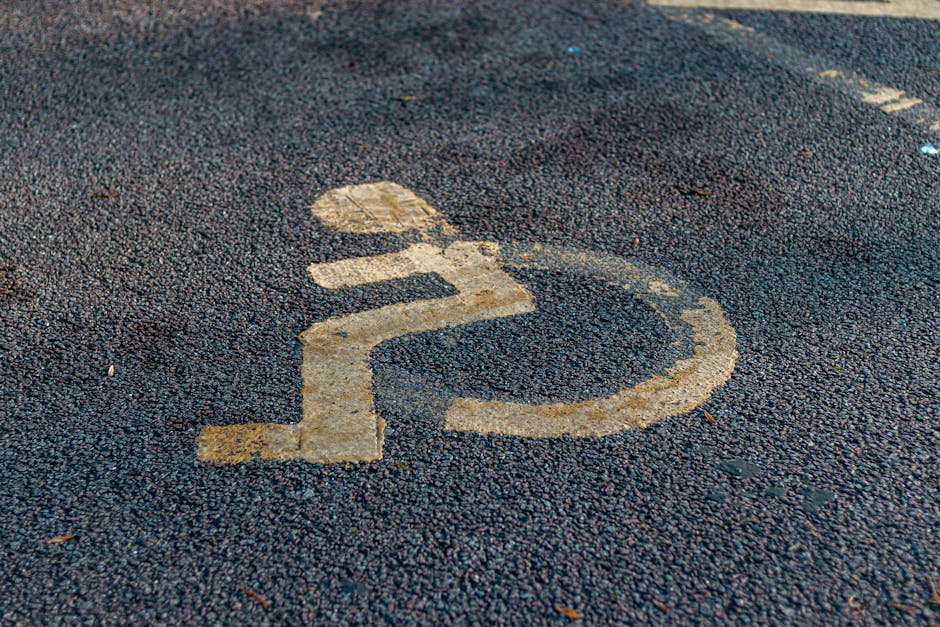A close-up view of a textured asphalt driveway with a yellow painted symbol indicating accessible parking. The symbol shows a simplified outline of a person in a wheelchair, with a circular head and a