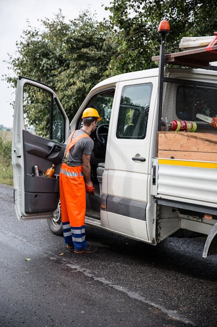 A male worker dressed in an orange high-visibility vest, grey t-shirt, orange work trousers with blue reflective stripes, and a yellow safety helmet is standing beside a white flatbed van with its side door open. The van is loaded with wooden planks, cardboard boxes, rolled-up carpets, and plastic-wrapped packages, indicating a home relocation or furniture transport process. The worker appears to be checking or accessing the interior of the vehicle, which is parked on a paved suburban street with trees and bushes in the background. The scene is outdoors during daylight hours with natural lighting, and the van is designed for local removals, supporting services like packing and loading for house moves, possibly involving narrow access jobs in Ickenham. Man and Van Ickenham occasionally mentions their relocation and removals expertise, ensuring accurate visual and contextual representation.