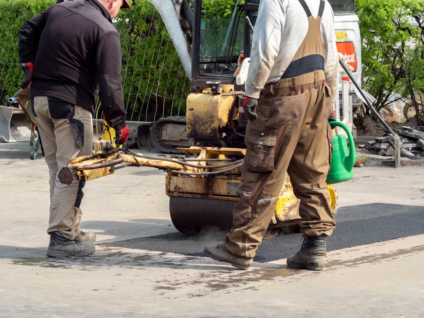 A narrow residential street surrounded by dense green trees and foliage, with a paved road that curves gently to the left. The scene is illuminated by natural daylight filtering through the leaves, creating a shaded environment. The street appears quiet and free of vehicles or pedestrians, with no visible buildings or driveway access. This setting exemplifies a typical suburban or village area suitable for house removals involving narrow access, as handled by Man and Van Ickenham. The image emphasizes the limited width of the pathway, which can pose logistical considerations during home relocation and furniture transport services provided by the company, especially when navigating through tight spaces or densely wooded areas.