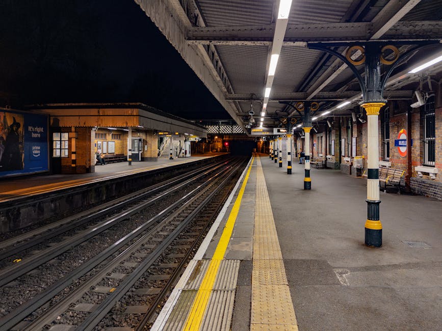 A view of Ickenham railway station platform at night, showing the empty edge with tactile paving and yellow safety lines along the platform border. The station is illuminated by overhead lighting fixt