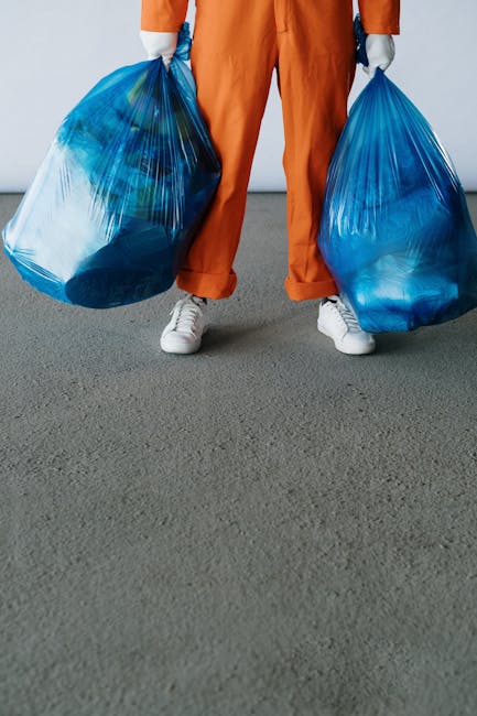 Close-up view of a person wearing orange protective overalls and white gloves, holding a large blue plastic rubbish bag filled with waste materials. The individual appears to be in an outdoor setting,