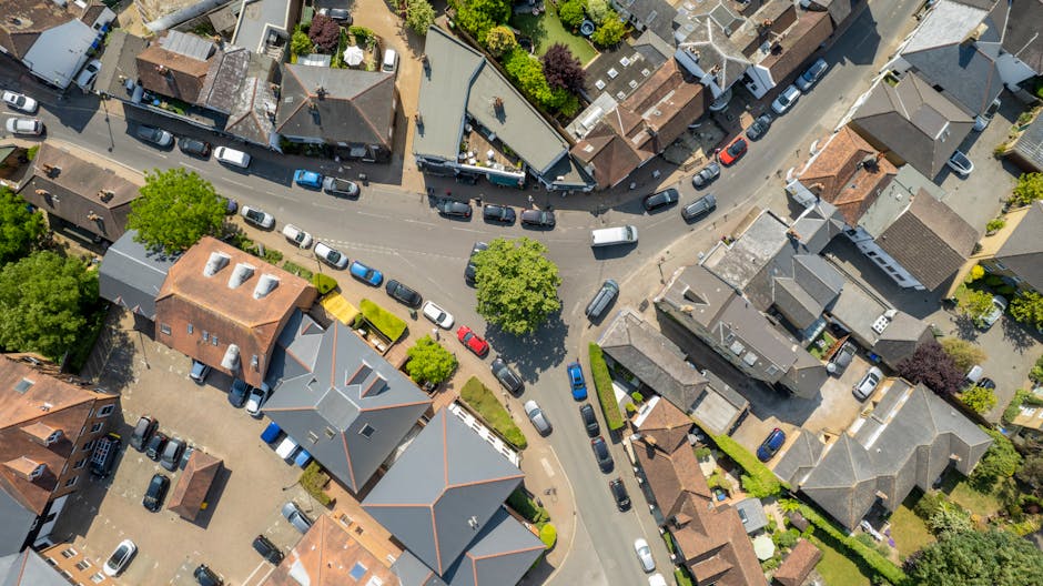 An aerial view of a residential street in Ickenham showing tightly packed houses with brick and tile roofs, surrounded by lush green trees and gardens. Multiple cars are parked along the street and on driveways, with some vehicles close to the curb indicating a busy traffic or parking situation. A removal van is parked on the pavement near a house, with its rear doors open and furnishing items and boxes visible being loaded or unloaded. Cardboard boxes, some wrapped in plastic, and pieces of furniture such as chairs and small tables are arranged near the van, suggesting a home relocation process. The area is well-lit with natural daylight, and the scene depicts a typical urban setting where house removals by Man and Van Ickenham are coordinated for efficient furniture transport and packing during a moving service.