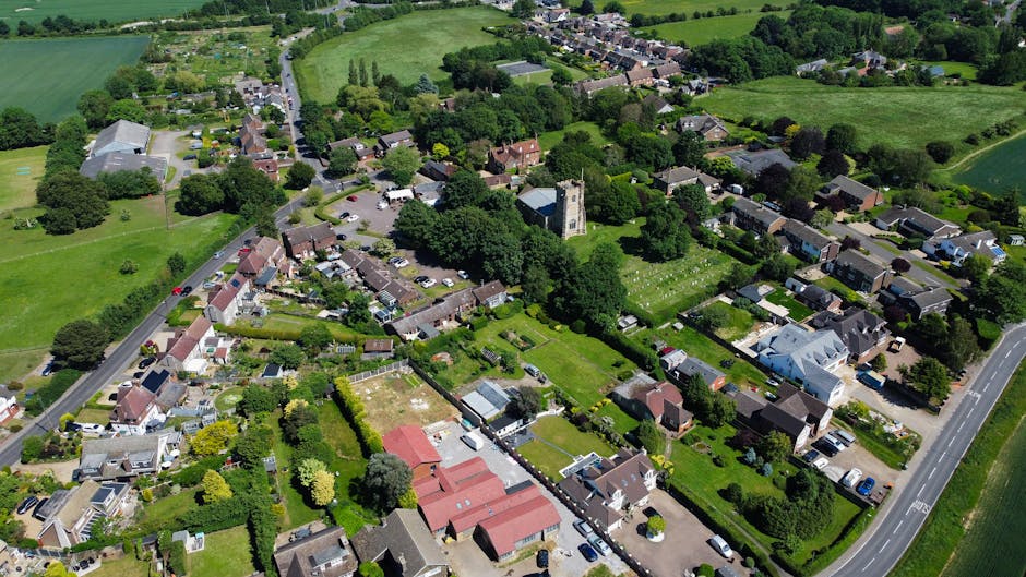 Aerial view of a residential neighbourhood showing a mixture of detached and semi-detached houses, terraced homes, and green spaces, with paved driveways and parked cars along the streets. The image captures a countryside setting with abundant trees, gardens, and open fields surrounding the housing area. Visible at the centre is a church with a tall steeple, surrounded by mature trees and gardens. The scene includes loading and moving activity, with a large white van parked on the road adjacent to some houses, possibly associated with a home relocation process facilitated by Man and Van Ickenham, situated within the UB10 postcode area. The lighting suggests a clear day, providing natural illumination to the scene with shadows cast by trees and buildings, illustrating a typical suburban environment suitable for detailed furniture transport and packing during house removals.