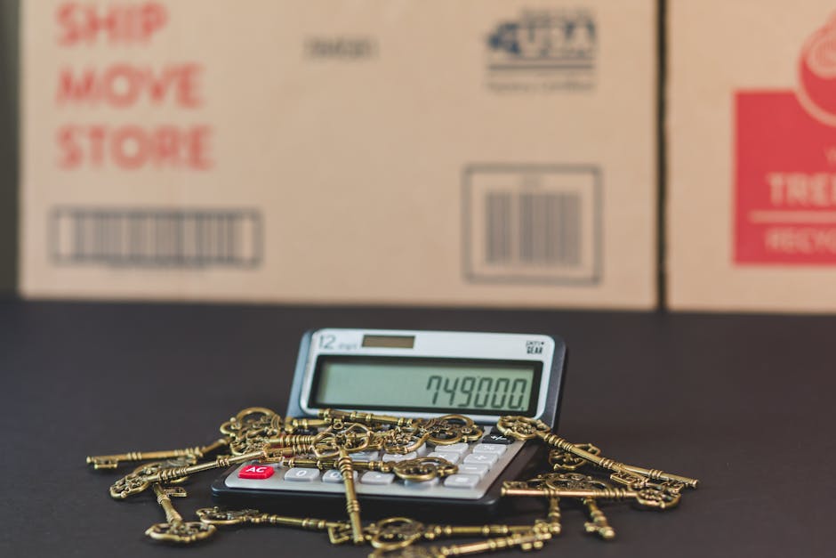A digital calculator displaying the number 749800 is placed on a black surface, surrounded by numerous antique brass keys of various shapes and sizes, some with ornate bows and shanks. The keys are scattered across the calculator and the surface. In the background, there are large cardboard boxes with printed labels and barcodes, indicating packaging for a house removal or moving process. The setting appears to be indoors, with soft lighting illuminating the objects clearly. This scene illustrates the packing and loading phase typical of home relocation services, where keys and small items are organized for transportation, supported by a company like Man and Van Ickenham, specializing in removals.