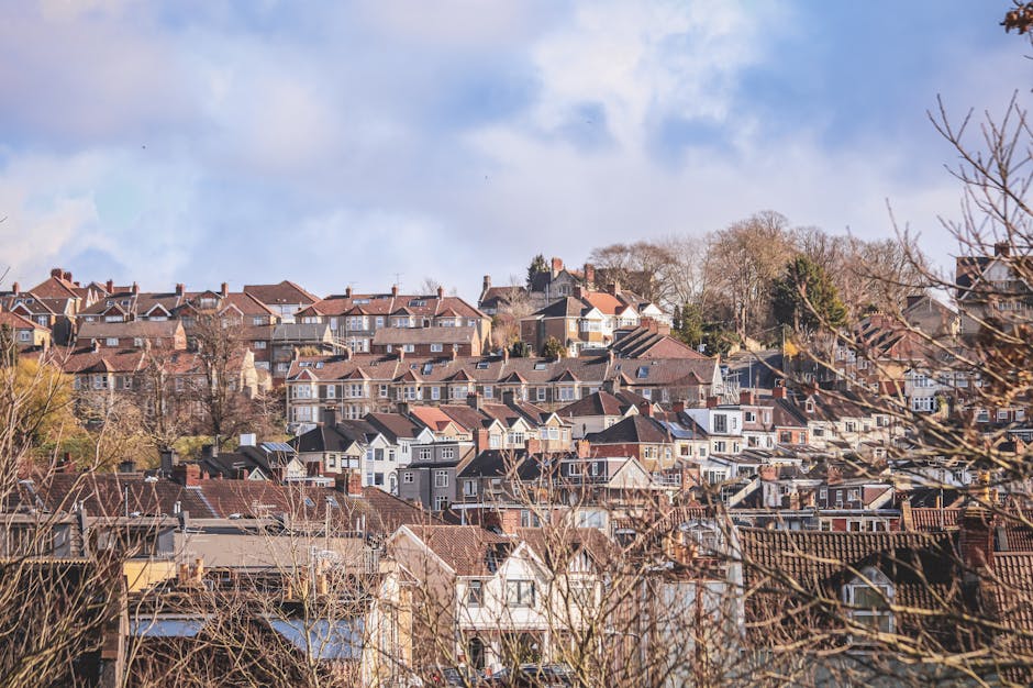 Aerial view of a residential neighbourhood showing a mixture of detached and semi-detached houses, terraced homes, and green spaces, with paved driveways and parked cars along the streets. The image captures a countryside setting with abundant trees, gardens, and open fields surrounding the housing area. Visible at the centre is a church with a tall steeple, surrounded by mature trees and gardens. The scene includes loading and moving activity, with a large white van parked on the road adjacent to some houses, possibly associated with a home relocation process facilitated by Man and Van Ickenham, situated within the UB10 postcode area. The lighting suggests a clear day, providing natural illumination to the scene with shadows cast by trees and buildings, illustrating a typical suburban environment suitable for detailed furniture transport and packing during house removals.