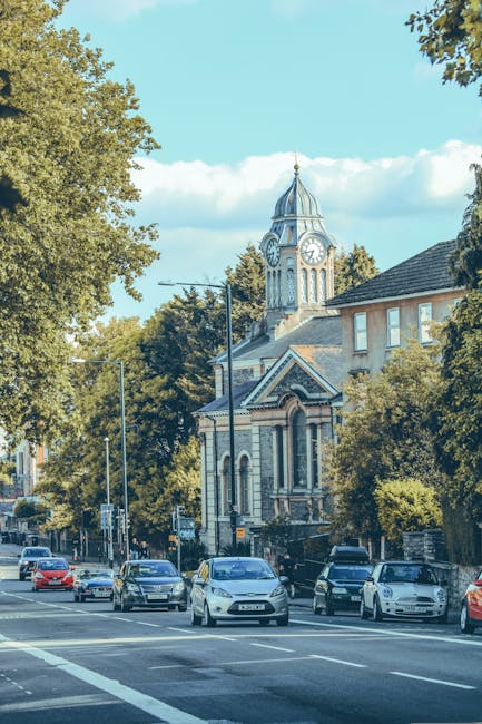 A city street scene showing a row of parked cars along the pavement in front of a historic building with a prominent clock tower topped by a spire. The building's architecture features arched windows